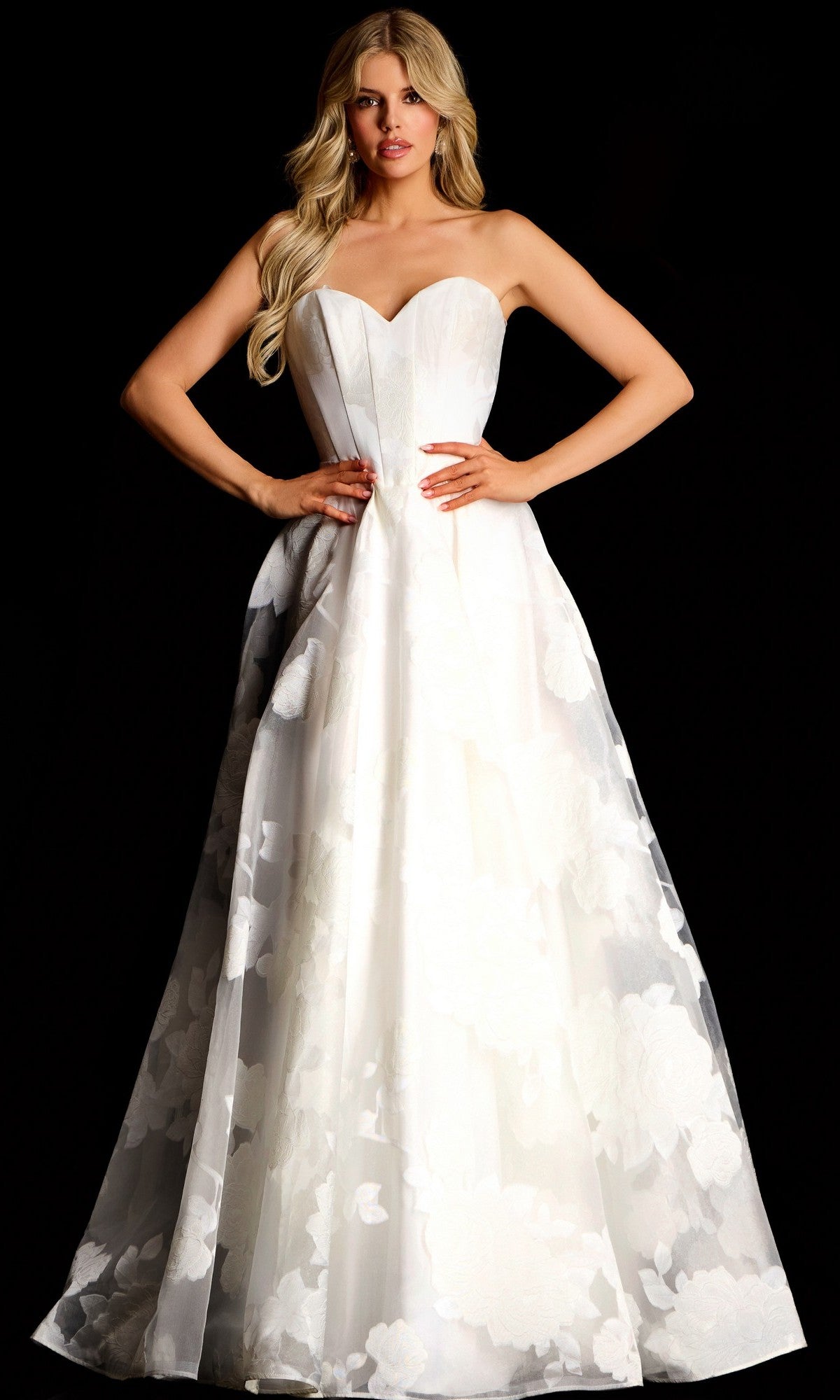 A woman in an off-white Jovani 47327 long prom dress with a corset bodice, strapless sweetheart neckline, and floral details stands against a black background.